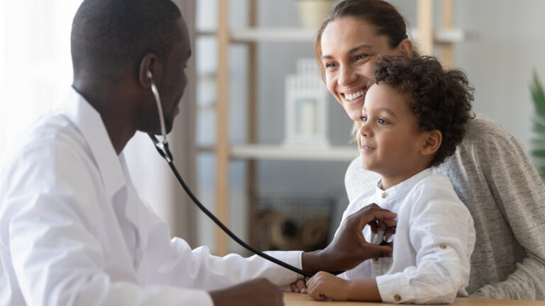 Um médico ouve o coração de um menino que está sentado no colo da mãe. Todos estão sorrindo