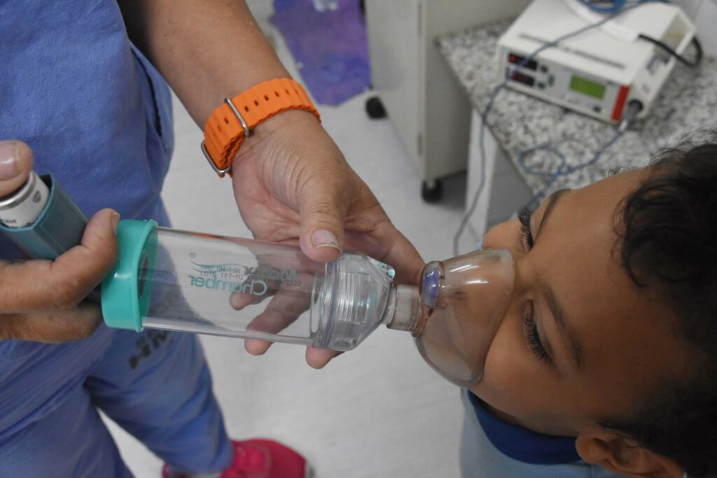 As mãos de uma profissional de saúde seguram um nebulizador sobre o rosto de um menino.
