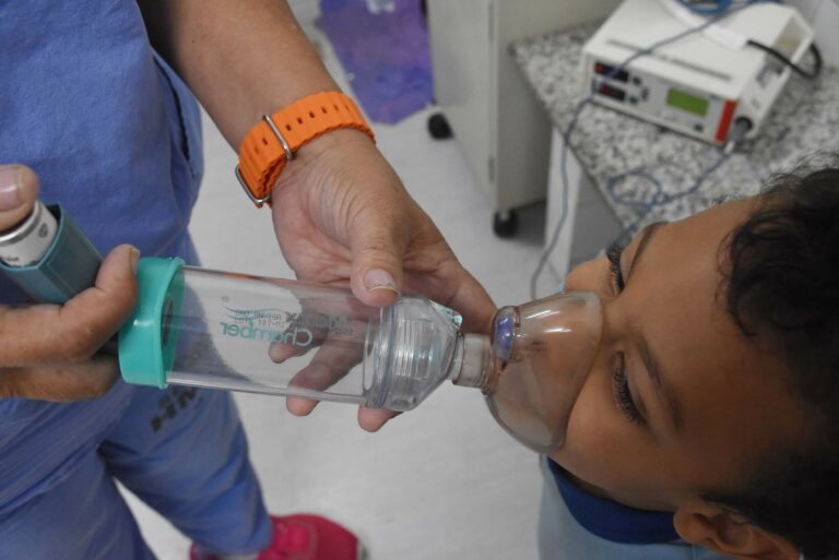 As mãos de uma profissional de saúde seguram um nebulizador sobre o rosto de um menino.