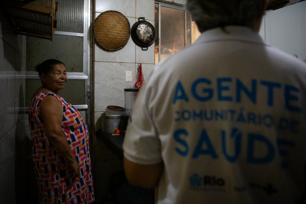 Uma pessoa está de costas, usando uma camiseta branca com a inscrição "agente comunitário de saúde" em azul. À sua frente, uma mulher idosa, vestindo um vestido colorido, sorri dentro de uma cozinha simples, sugerindo uma visita domiciliar.
