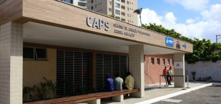 Fachada de um Centro de Atenção Psicossocial (CAPS) identificado como "Jardim Armação". Na frente do local há um longo banco de madeira onde algumas pessoas estão sentadas.