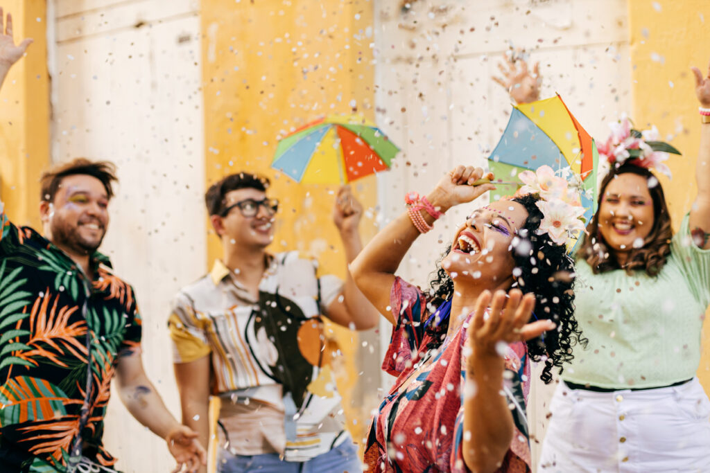 Grupo de amigos comemorando o Carnaval ao ar livre, jogando confetes no ar em clima de diversão. Eles usam roupas coloridas, acessórios festivos e seguram sombrinhas de frevo.
