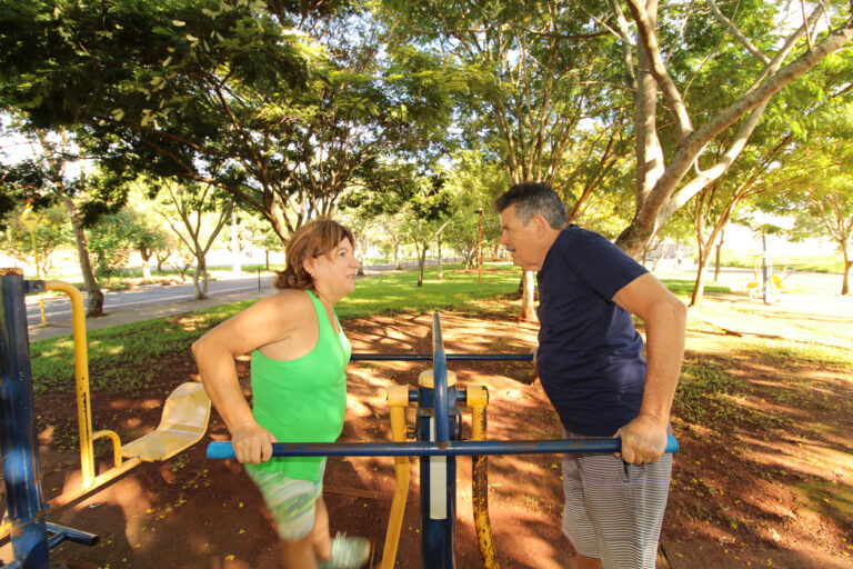 Uma mulher e um homem se exercitam em uma academia ao ar livre em um parque arborizado. Eles vestem roupas esportivas e utilizam um equipamento de barras paralelas para alongamento ou flexões.