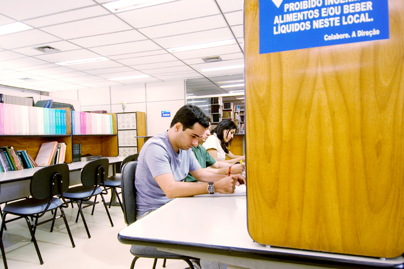 Dois homens e uma mulher trabalham atentamente, sentados lado a lado.. Atrás deles, uma bancada com livros e cadeiras vazias. Ao fundo, estantes com livros.