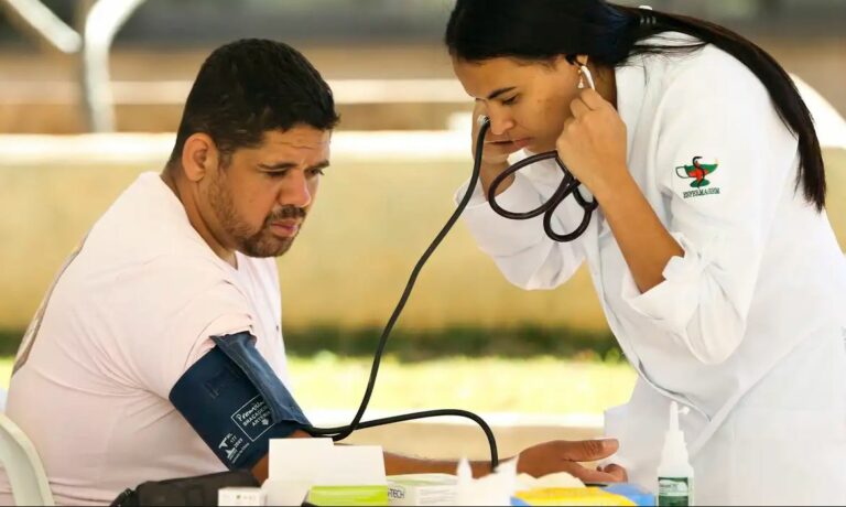Um homem está sentado com um equipamento de medir pressão arterial em um de seus braços, enquanto uma mulher está curvada sobre ele, olhando para as informações deste equipamento.